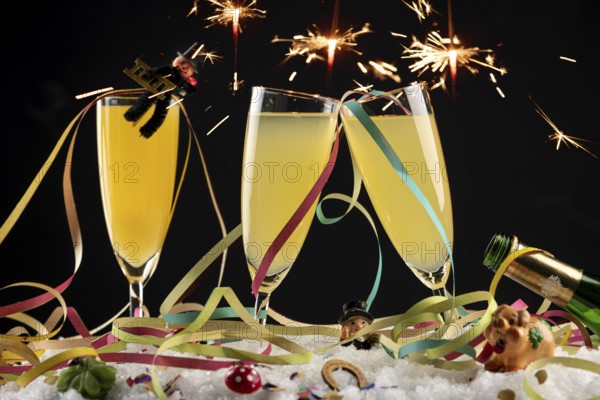 Sparks fly over three champagne glasses with lucky New Year's charms and streamers in the snow