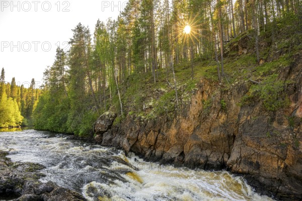 A river flows through a thick forest as the sun shines over the treetops and illuminates the rocks, Hossa National Park, Juuma, Kuusamo, Northern Ostrobothnia region, Finland