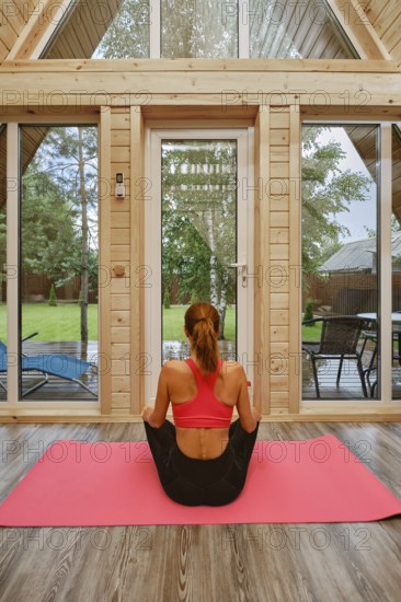 Back view of a woman sitting cross-legged on a pink yoga mat inside a wooden cabin. Outside, there is a green garden with chairs and sunlight coming through the trees
