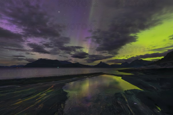 The Vestfjord in Nordland, Norway, has an enchanting atmosphere at night. Dark clouds and vivid aurora, polar lights dance over the fjord. The rugged cliffs and majestic mountains are reflected in the calm water and create a mystical backdrop