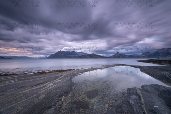 The Vestfjord in Nordland, Norway, offers a dramatic atmosphere near Bodø. Dark clouds are illuminated by the setting sun in shades of red and yellow. The rugged cliffs and mountains are reflected in the quiet fjord, which offers a breathtaking view of Lofoten