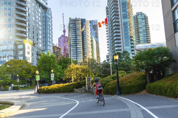 Woman in a helmet and backpack riding a bicycle on a paved path in vancouver, british columbia, surrounded by modern buildings, green trees, and a clear sky