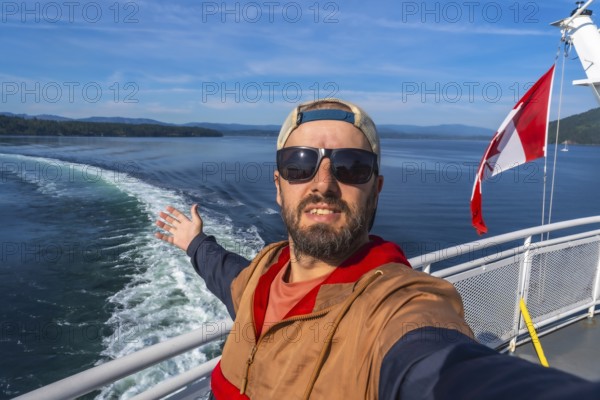 Tourist wearing sunglasses and a cap enjoys ferry trip from victoria, vancouver island, to vancouver, british columbia, opening his arms and showing canadian flag and ocean wake