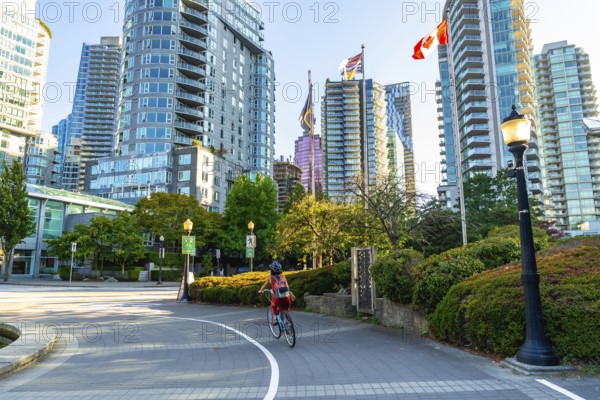 Student wearing a helmet and backpack cycling on a bike lane in downtown vancouver, surrounded by modern buildings, green spaces, and canadian flags