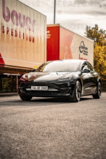 Black car in front of a truck sign on a sunny road in autumn, Deer E-Carsharing, Tesla Model 3, Calw, Germany