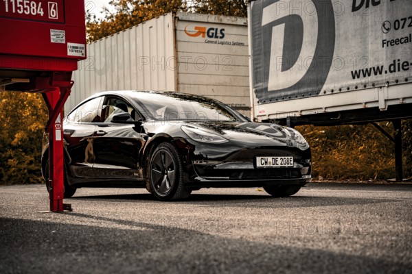 Black car between trucks in a parking lot with autumn scenery, Deer e-car sharing, Tesla Model 3, Calw, Germany