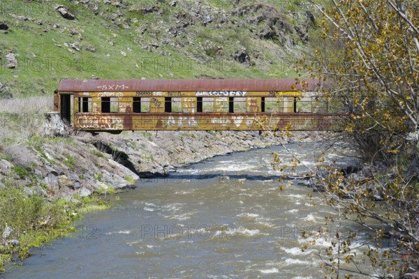 A rusted train car with graffiti stands over a flowing river in a green, hilly landscape, discarded railroad car used as a footbridge, Paravani river, Akhalkalaki, Akhalkalaki, Samtske-Javakheti region, Samtske Javacheti, Lesser Caucasus, Georgia