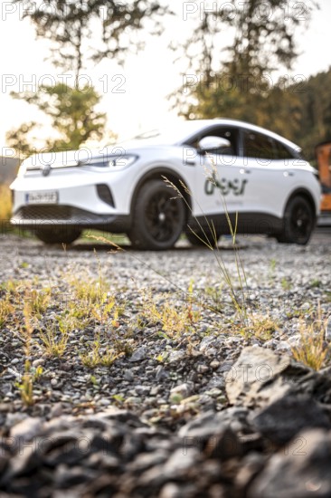 White car photographed from a blurred perspective on rocky ground with plants in the foreground, VW ID4 electric car, Deer Carsharing, Calw, Germany