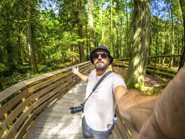 Photographer with hat and sunglasses taking a selfie while walking on a wooden path inside the amazing cathedral grove forest during a sunny summer day on vancouver island, british columbia, canada