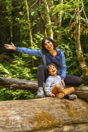 Cheerful mother and her son are sitting on a large fallen log in cathedral grove forest on vancouver island, british columbia, canada, enjoying the beauty of nature during summer