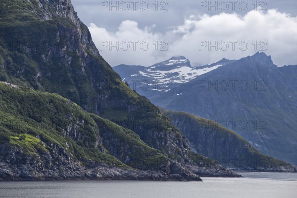 Green hills and snow-capped mountains overlook a calm sea as thick clouds cover the sky, Norway
