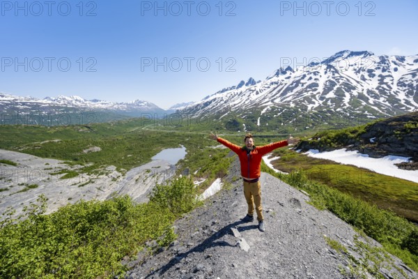 Hiker stretches his arms in the air, on the glacial moraine, view over the wide valley of the Tsina River with mountains, Worthington Glacier State Recreational Site, Chugach Mountains, Alaska, USA