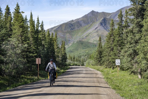 Young woman riding a bicycle on a dirt road through taiga, mountainous landscape, Denali Park Road, Denali National Park and Preserve, Alaska, USA