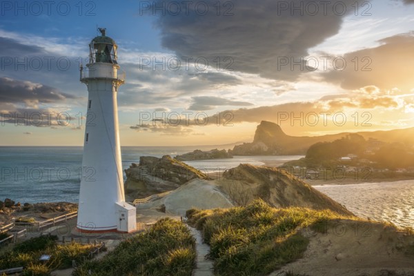The Castlepoint lighthouse on a rock, ocean, Deliverance Cove and Castle Rock in the background. Evening, golden hour, sunset. Castlepoint, Wairarapa Coast, Wellington Region, North Island, New Zealand
