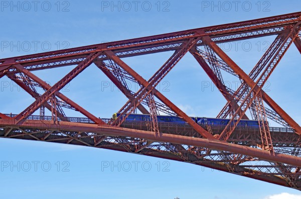 A train crosses a massive red steel bridge under a blue sky, Firth of Forth, Edinburgh, Scotland, United Kingdom