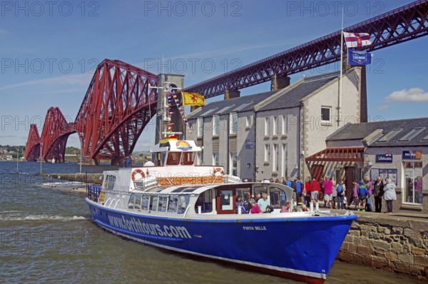 A tourist boat in front of a distinctive red bridge structure next to a building, railway bridge, Firth of Forth, Edinburgh, Scotland, Great Britain
