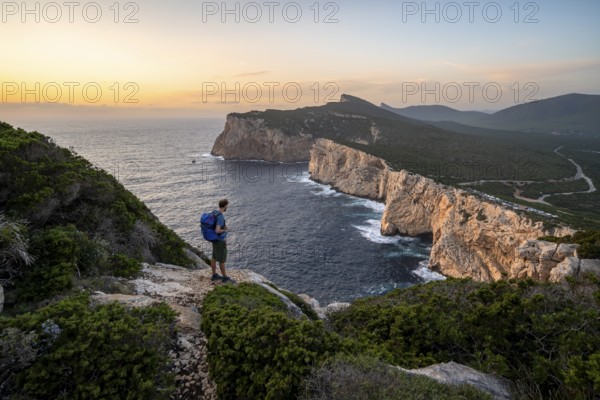 Tourist in front of coastal landscape at sunset, steep cliffs by the sea, cliffs in the evening light, Capo Caccia, Alghero, Sardinia