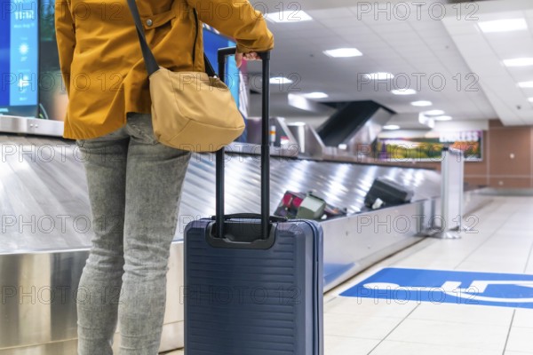 Tourist waiting at the baggage claim area in an airport, holding a suitcase while observing the conveyor belt for arriving luggage after a long journey