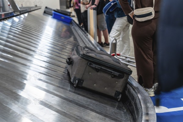 Passengers waiting at the baggage claim carousel in the airport, watching as a suitcase arrives on the conveyor belt. The atmosphere buzzes with anticipation and travel excitement