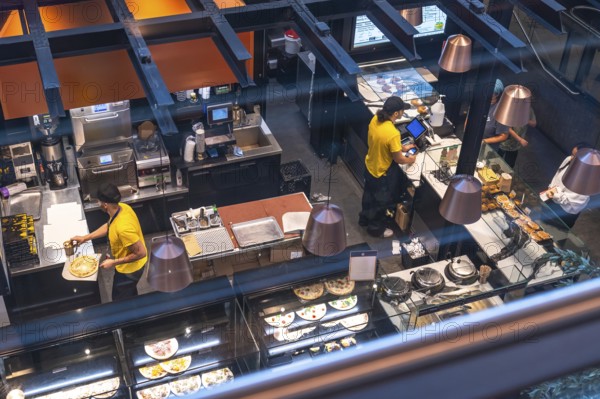 Chefs working in a busy airport restaurant kitchen prepare various dishes, including pizza and pastries, for travelers in a fast paced culinary environment
