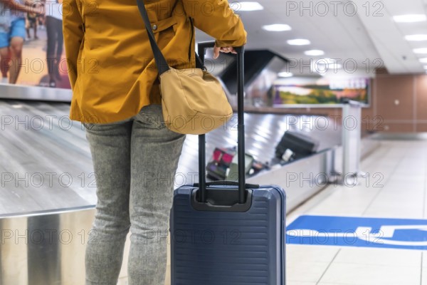 Tourist is waiting for luggage at baggage claim carousel in airport terminal after arrival, holding suitcase and shoulder bag, travel and tourism concept