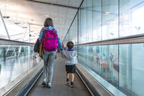Mother and son walking hand in hand on a moving walkway in a modern airport terminal, sharing a special moment as they embark on their journey together, filled with excitement and love