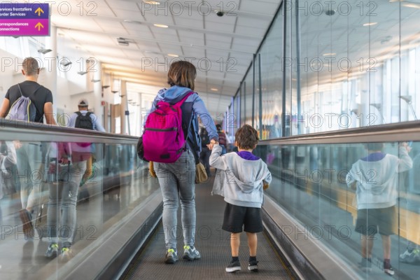 Mother and son with backpacks holding hands walking on a moving walkway in a modern airport terminal, heading towards border services and connecting flights, surrounded by other passengers