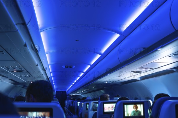 Airplane cabin illuminated with blue ambient light, passengers sitting on their seats, some watching movies on the screens embedded in the headrests