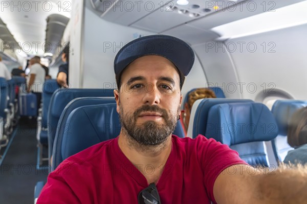 Bearded man capturing a selfie while seated in an airplane during a flight, with fellow passengers boarding in the background, embodying the excitement of travel and adventure