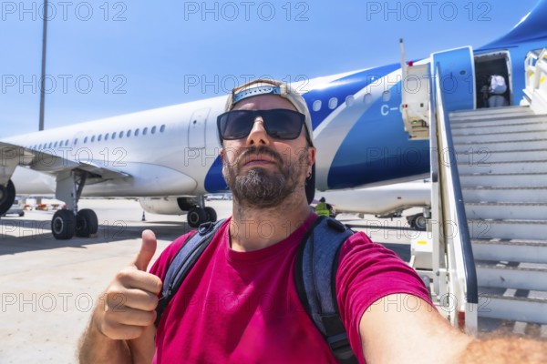 Happy bearded tourist wearing sunglasses and backpack taking selfie showing thumbs up gesture while boarding airplane at airport on sunny summer day