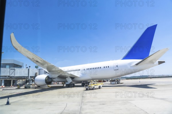 White and blue passenger airplane parked at the airport gate, getting ready for departure under a bright sunny sky, with clear blue horizons highlighting a perfect travel day