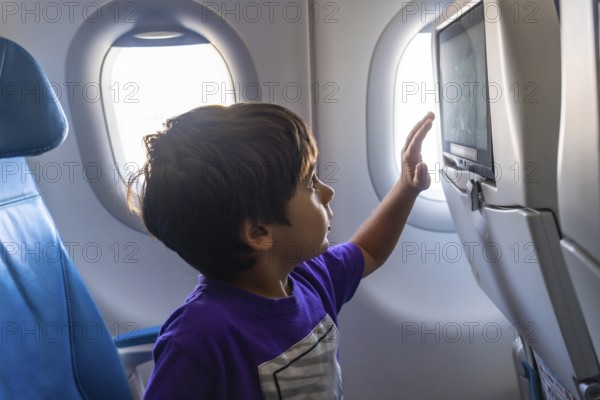 Young boy enjoying his first flight, gazing out the window with wonder and touching the screen on the back of the seat in front of him, experiencing the thrill of air travel