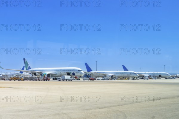 Several airplanes are parked on the tarmac at an airport, waiting for their next flights under a clear blue sky, with ground crew vehicles attending to them