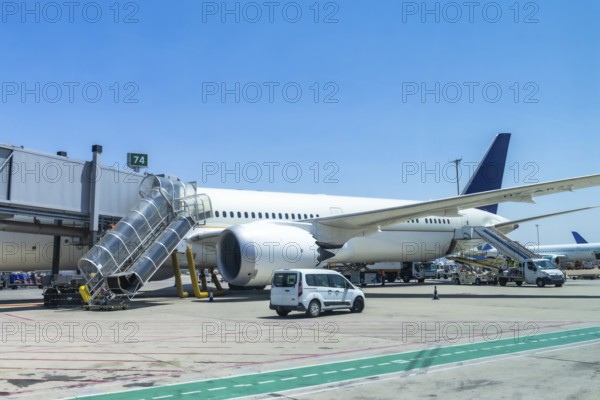 Modern passenger airplane parked at airport gate, connected to jet bridge, with ground crew and support vehicles preparing for passenger boarding on a sunny day