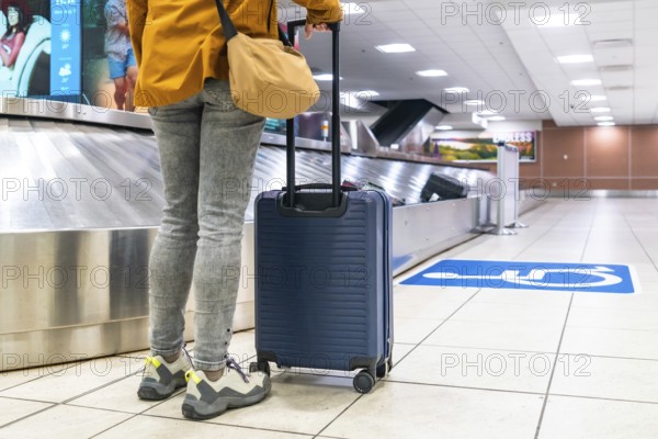 Traveler waiting by the baggage claim carousel at the airport, holding a trolley suitcase and a shoulder bag, anticipating the arrival of luggage after a long journey