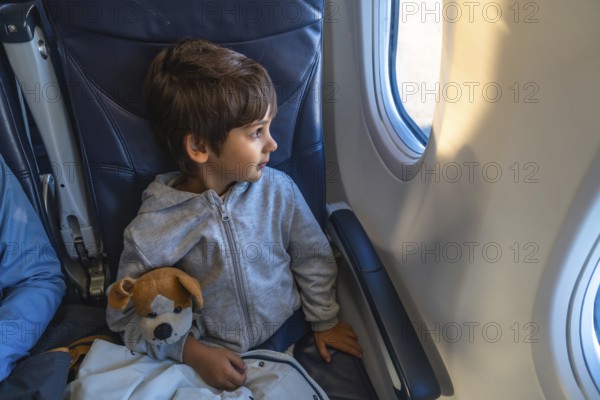 Curious child traveling by plane, clutching a plush toy while gazing out the window, marveling at the expansive sky and fluffy clouds during an exciting flight adventure