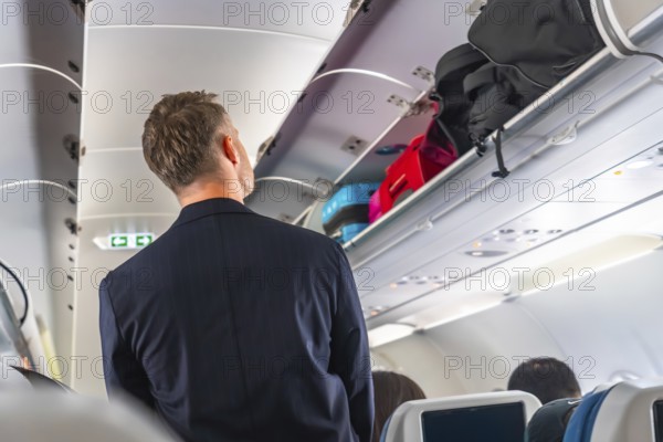 Businessman searching for space to store carry on luggage in the overhead compartment of a crowded passenger airplane during a busy flight, navigating the challenges of air travel