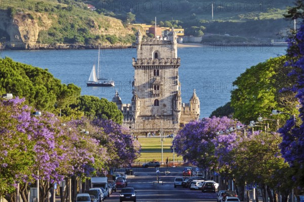 Scenic view of Belem Tower in Lisbon, Portugal, seen over a street with blooming purple jacaranda flower trees street with tourist sailboats on the Tagus River on sunset. Portugal
