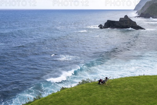 Woman sitting comfortably in deckchair in a meadow and watching waves in the sea, Madeira, Portugal