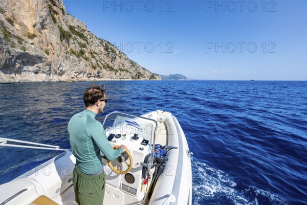 Young man rides a motorboat along the picturesque rocky coast, cliffs and blue sea, Golfo di Orosei, Baunei, Sardinia, Italy