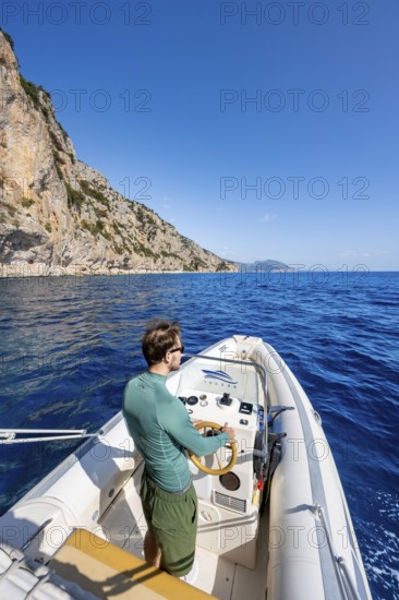 Young man rides a motorboat along the picturesque rocky coast, cliffs and blue sea, Golfo di Orosei, Baunei, Sardinia, Italy