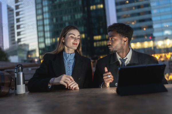 Business colleagues of different ethnicities are sitting at a table with a tablet, engaging in a focused discussion outdoors against the backdrop of modern city buildings at dusk