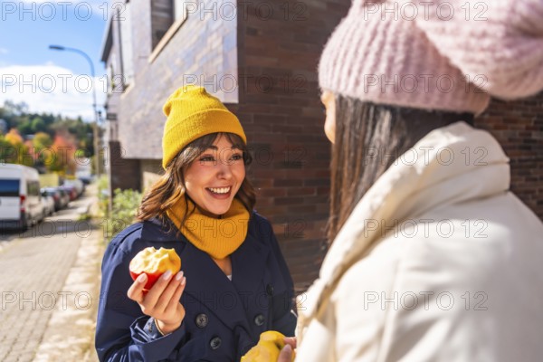 Two cheerful women friends enjoying fresh apples and engaging in a conversation outdoors on an urban street during a bright autumn day, reflecting healthy eating and friendship
