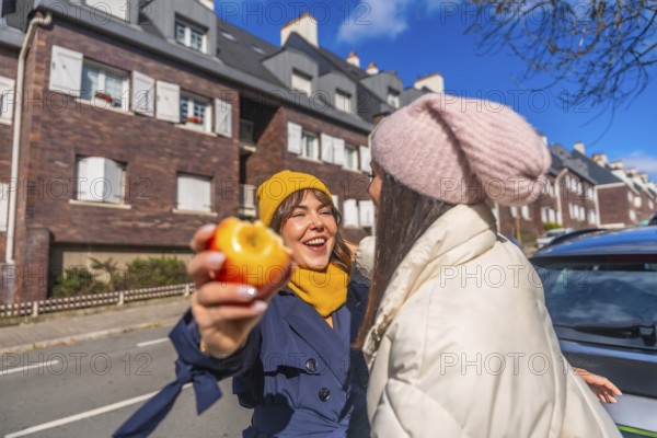 Two young women laughing and sharing a healthy snack on a sunny urban street, bundled in coats and beanies, enjoying friendship, conversation and carefree city life