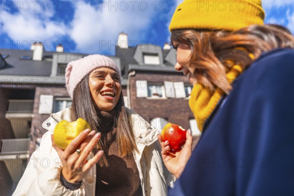 Two cheerful women friends enjoying a healthy apple snack and talking outdoors on a sunny winter day, wearing warm clothing and hats with an urban building background
