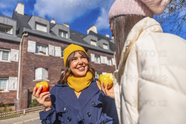 Two young women laugh and chat on a sunny autumn city street, sharing fresh apples and cozying up in beanies and coats while enjoying a healthy, carefree moment together