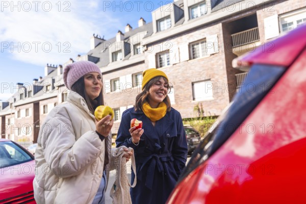 Two young adult women friends enjoying a healthy snack of apples while walking on a city street, embracing a sustainable and active urban lifestyle in a cheerful moment