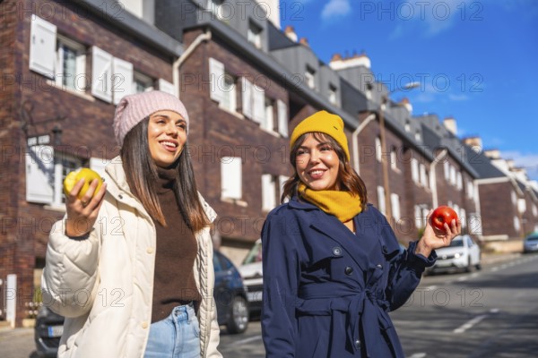 Two smiling young women friends are enjoying a sunny winter day, walking along a city street and holding fresh apples, promoting healthy eating and an active urban lifestyle