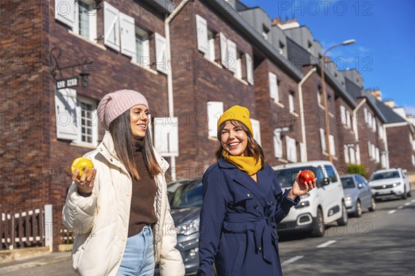 Two cheerful young women enjoying a healthy snack of apples while walking and smiling on a city street during autumn, embracing a vibrant urban lifestyle
