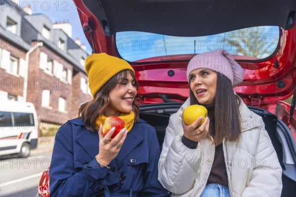 Two cheerful women sit in a car trunk on a winter road trip, sharing apples, laughing and talking during a cozy healthy snack break, bonding over fresh fruit and adventure